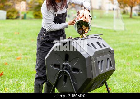 Una donna sta scaricando un piccolo bidone di scarti di cucina in un composter tumbling esterno nel giardino del cortile. Queste unità in plastica con gambe in metallo possono ruotare a. Foto Stock