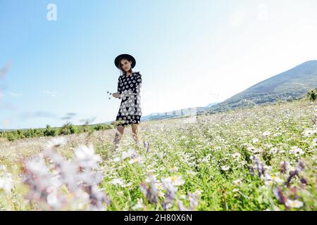 una bella donna in un abito e cappello che cammina attraverso un campo di margherite Foto Stock