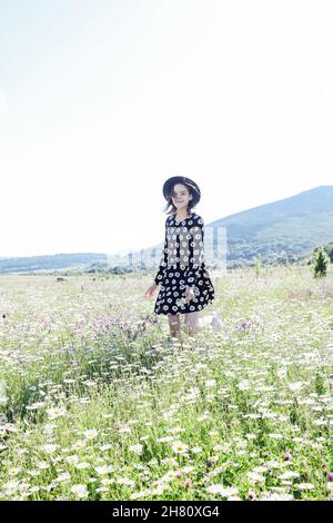 una bella donna in un abito e cappello che cammina attraverso un campo di margherite Foto Stock