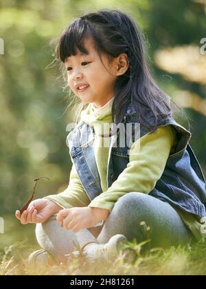 ritratto all'aperto di una bambina asiatica seduta su erba felice e sorridente Foto Stock