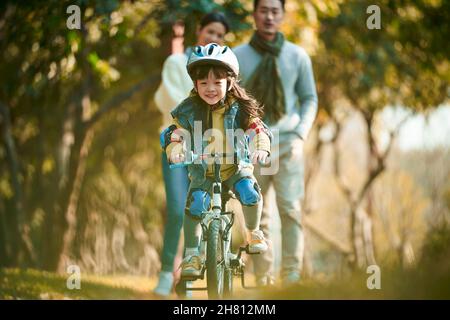 bambina asiatica con casco e completo di protezione in bicicletta nel parco della città con i genitori che guardano da dietro Foto Stock