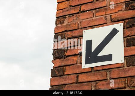 Freccia nera su sfondo bianco rivolta verso il basso e verso sinistra. Sullo sfondo di mattoni rossi e cielo blu. Spazio di copia. Foto Stock