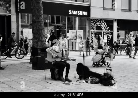 Cardiff, Galles, agosto 18 2021: Immagine monocromatica di un giovane uomo d'affari siede in Queen St. Cardiff suona la sua chitarra per gli acquirenti. Foto Stock