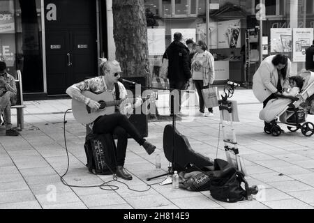 Cardiff, Galles, agosto 18 2021: Immagine monocromatica di un giovane uomo d'affari siede in Queen St. Cardiff suona la sua chitarra per gli acquirenti. Foto Stock