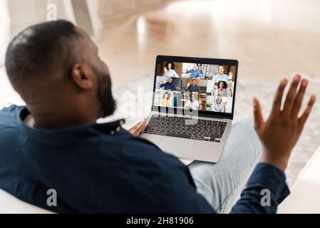 Incontro mattutino con colleghi diversi. Tipo afroamericano che utilizza l'app per computer per la comunicazione virtuale con un gruppo di persone internazionali, per videochiamare, salutare, stare a casa, guardare la schiena Foto Stock