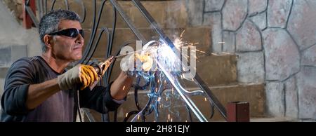 Uomo che indossa occhiali protettivi salda metallo con saldatrice in casa privata. Le scintille provenienti dalla saldatura volano sui lati. Banner. Foto Stock