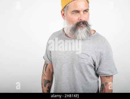 Ritratto di un uomo dai capelli grigi, bearded, tatuato in testa gialla e t-shirt grigia che posa isolata in uno studio su sfondo bianco. Foto Stock