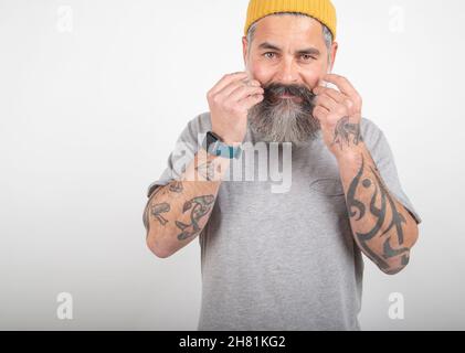 Ritratto di un uomo grigio pelato, bearded, tatuato in t-shirt grigia posando isolato in uno studio a contatto dei suoi baffi. Foto Stock