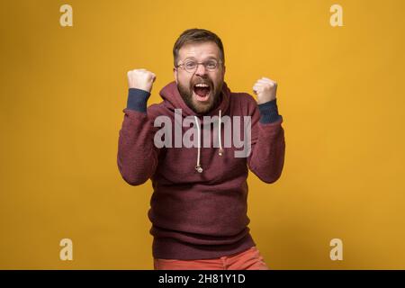 L'uomo caucasico felice fa un gesto vincitore, egli innalza energicamente i pugni e grida di gioia. Sfondo giallo. Foto Stock