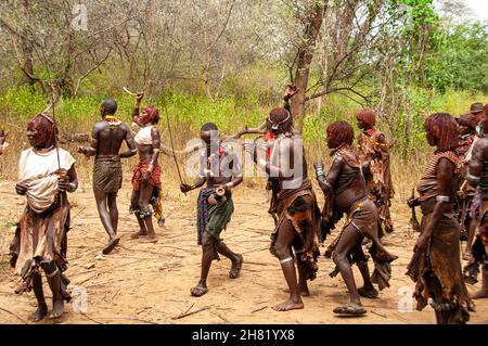 Uomini e donne che ballano e cantano prima della cerimonia di salto del toro per la tribù di Hamar in Etiopia, donne e uomini tengono rami lunghi e sottili usati per montare il Foto Stock
