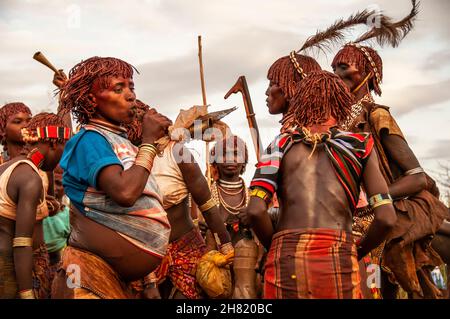 Le donne della tribù di Hamar cantano, ballano e soffiano corna per fare rumore prima dell'inizio della cerimonia di salto del toro Foto Stock