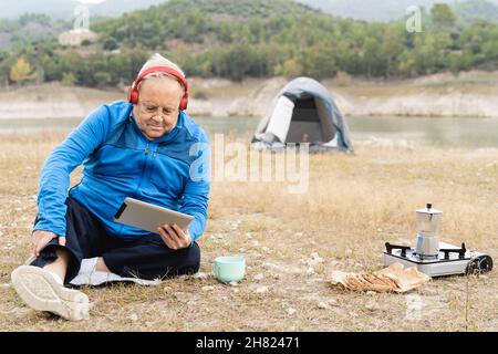 Uomo anziano campeggio con tenda durante la lettura libro e bevande caffè con lago sullo sfondo - persone anziane viaggio Foto Stock