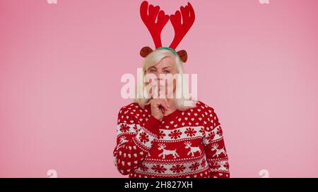 Anziana donna nel maglione di Natale preme indice dito a labbra rende segreto gesto silenzio Foto Stock