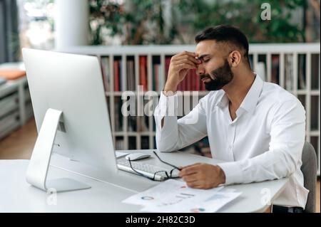 Stanco uomo indiano esausto, proprietario dell'azienda o dipendente dell'ufficio, si siede alla scrivania con gli occhi chiusi, gli sfrega gli occhi, la sensazione di mal di testa, lo stress e la fatica da lavoro straordinario, hanno bisogno di riposo e rottura Foto Stock