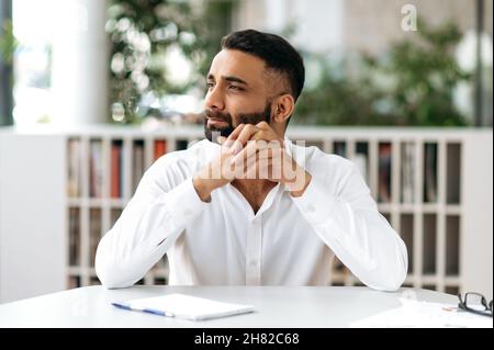 Uomo indiano concentrato, uomo d'affari, manager, indossando una camicia bianca, seduto al tavolo nel suo ufficio, guardando penosamente a lato, pensando a un nuovo progetto o sognando una vacanza Foto Stock