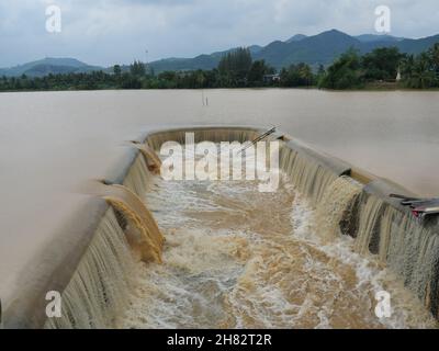 Acqua marrone nella diga che trabocca nel canale di fuoriuscita con montagna e foresta verde sullo sfondo, inondazione nella stagione delle piogge, Thailandia Foto Stock