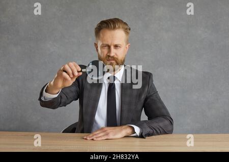 Serio uomo d'affari concentrato seduto ad un tavolo e guardando attraverso una lente d'ingrandimento. Foto Stock