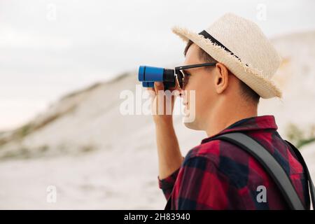 Viaggiatore maschile con uno zaino che guarda attraverso binocoli all'aperto nella natura. Il ragazzo guarda attraverso binocoli sulla collina. Un uomo con binocolo e un Foto Stock