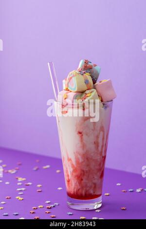 Tazza di vetro con milkshake e sciroppo di frutta con confetti di stelle, marshmallow multicolore e paillette di vetro su sfondo viola brillante Foto Stock