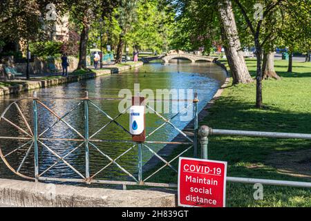 Un segno sociale di distanziamento e sanitizzante mano accanto al fiume Windrush nel villaggio Cotswold di Bourton sull'acqua, Gloucestershire Regno Unito durante il C. Foto Stock