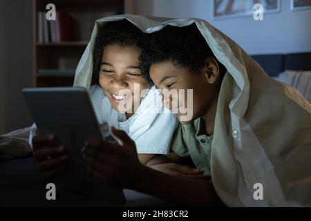 Felici bambini afroamericani sdraiati sotto il piumone e leggendo online libro su tablet in camera oscura Foto Stock