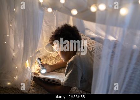 Curioso ragazzo nero con capelli ricci che giacciono all'interno della tenda da gioco decorata con luci di Natale e libro di lettura con torcia Foto Stock