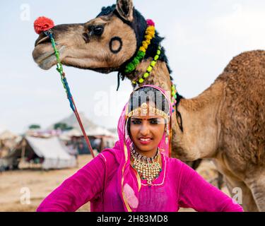 Giovane donna zingara in abiti colorati e gioielleria brillante si erge prossimo cammello decorato in vendita a Pushkar, India. Foto Stock