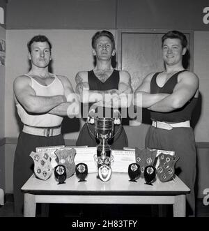 1965, storico, tre membri della squadra di sollevamento pesi del servizio civile degli uomini, le braccia piegate, posano per una foto da un tavolo che mostra il loro trofeo vincente, il Perth Tophy, più varie targhe di vittoria e il campionato diplomi, Fife, Scozia, Regno Unito. Foto Stock