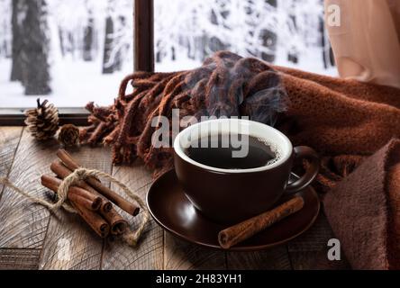 Una tazza di caffè fumante su un tavolo di legno vicino a una finestra con sfondo invernale Foto Stock