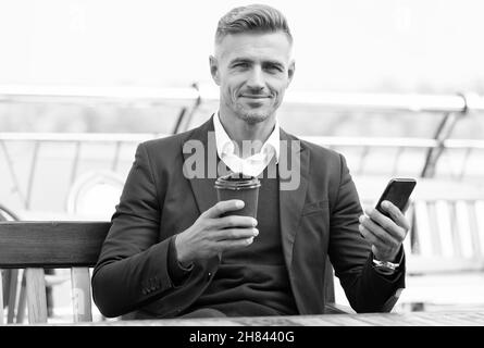 Il caffè mi completa. Il boss tiene la tazza da asporto e lo smartphone. Pausa caffè all'aperto. Bevanda calda. Tecnologia moderna. Colazione al mattino. Affari Foto Stock