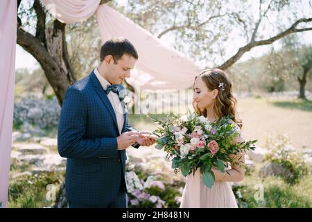 Lo sposo mette un anello sul dito della sposa con un bouquet nel parco Foto Stock