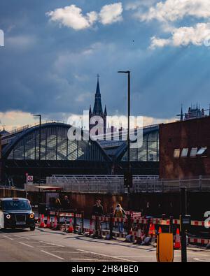 Shot of Kings Cross station, Londra, dal retro in estate sotto i cieli estivi con raggi di luce dal sole come un taxi nero guida e la gente a piedi. Foto Stock