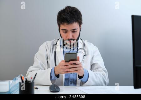 Giovane medico che utilizza lo smartphone seduto alla scrivania in un ufficio medico. Foto Stock