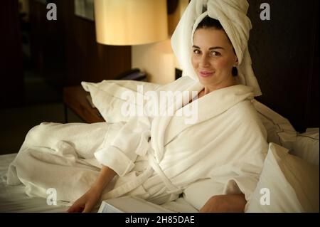 Bella donna europea di 30 anni dai capelli scuri in accappatoio bianco e la testa avvolta in un asciugamano, rilassante dopo il bagno, sdraiata su un letto Foto Stock