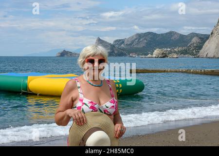 Ritratto di donna anziana che si trova in piedi sulla spiaggia di ciottoli con cappello di paglia nelle sue mani su onde surf e sfondo scogliera a Sudak, Crimea, Russia. Foto Stock