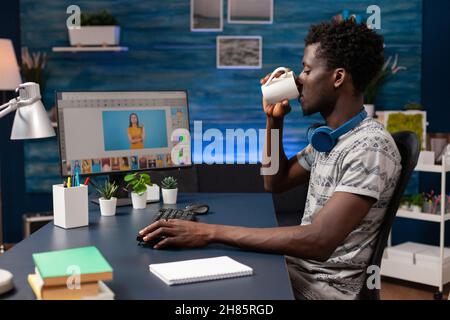 Fotografo afroamericano che beve caffè mentre si modifica la fotografia digitale utilizzando il software di editing post-produzione. Giovane illustratore editor lavorando in remoto alle foto ritoccare in studio creatività Foto Stock