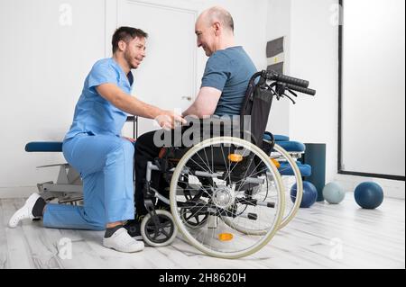Fisioterapista maschile che aiuta un paziente con una disabilità che utilizza una sedia a rotelle per alzarsi in ospedale di riabilitazione. Foto di alta qualità. Foto Stock