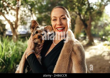 Felice donna elegante sorridendo alla macchina fotografica mentre tiene il suo cucciolo all'aperto. Donna di fascia alta di età media in piedi da sola in un parco durante il giorno. Femmina Foto Stock