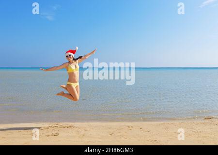 donna felice a santa cappello e occhiali da sole che saltano sulla spiaggia di sabbia Foto Stock