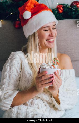 Donna sorridente bionda che indossa abito in maglia bianca Santa Hat sedersi a letto in camera da letto decorata a Natale e bere cioccolata calda con marshmallows Foto Stock