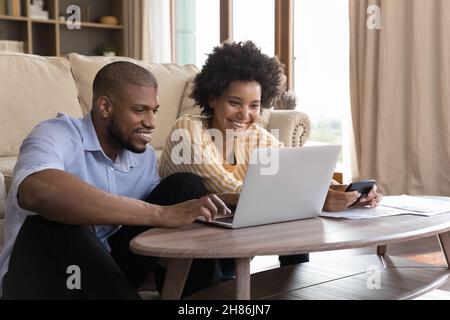 Felice giovane coppia nera che usa il laptop, guarda lo schermo, sorridente Foto Stock
