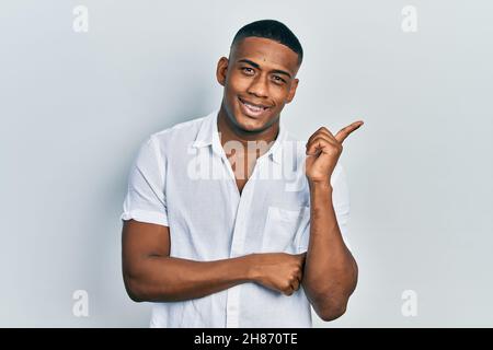 Giovane uomo nero che indossa una camicia casual bianca con un grande sorriso sul viso, puntando con mano e dito verso il lato guardando la fotocamera. Foto Stock