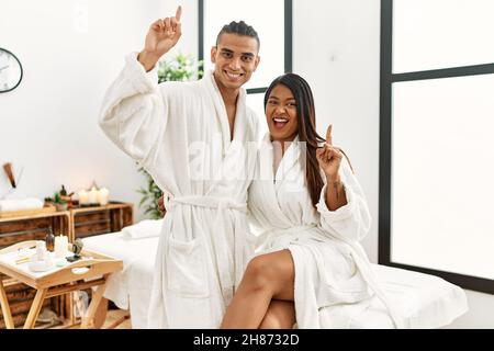 Giovane coppia latina che indossa un asciugamano in piedi al centro di bellezza puntando il dito verso l'alto con un'idea di successo. Uscito e felice. Numero uno. Foto Stock