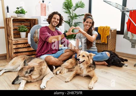 Giovane coppia ispanica che fa la lavanderia con i cani sorridenti nell'amore che fa la figura del simbolo del cuore con le mani. Concetto romantico. Foto Stock