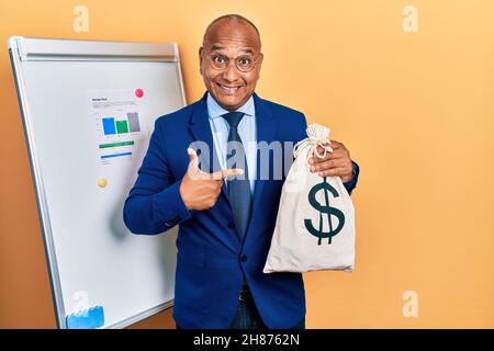 Uomo latino di mezza età che indossa tuta da lavoro che tiene dollari borsa sorridente felice indicando con mano e dito Foto Stock