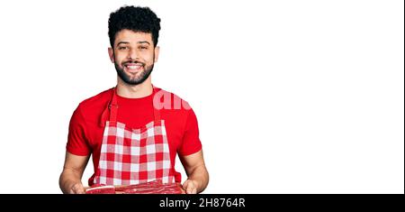 Giovane uomo arabo con la barba che tiene la tavola con carne cruda sorridendo con un sorriso felice e fresco sul viso. Mostrando i denti. Foto Stock