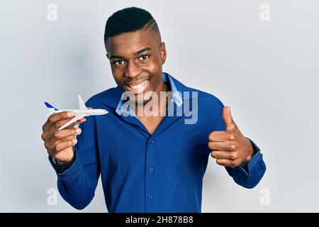 Giovane afroamericano uomo che tiene il giocattolo dell'aeroplano sorridente felice e positivo, pollice in su che fa eccellente e segno di approvazione Foto Stock