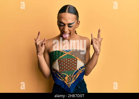 Uomo ispanico che indossa make up e lunghi capelli che indossa i woman top gridando con pazza espressione facendo rock simbolo con le mani in su. Star della musica. Pesante concep Foto Stock