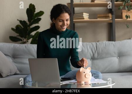 Happy casual latino femmina goccia moneta a piggybank a casa Foto Stock