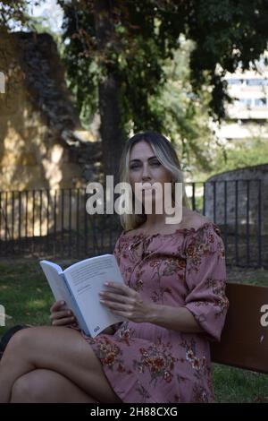 donna che legge un libro in un parco durante la giornata di sole guardando la macchina fotografica, guardando un libro, seduto su una panca, durante un inverno, giorno d'estate Foto Stock